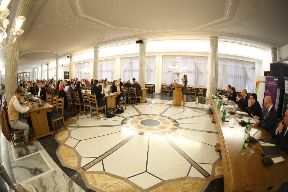 Na środku dużej sali znajduje się drewniana mównica, przy której stoi kobieta. Przemawia ona w stronę publiczności, która siedzi przy trzech bardzo długich drewnianych stołach ustawionych do siebie równolegle. Stoły są ustawione wzdłuż ozdobnych filarów. Na podłodze widnieje ornament w formie rozety. Za mównicą ustawiony jest długi drewniany stół przy którym zasiadają członkowie komisji.